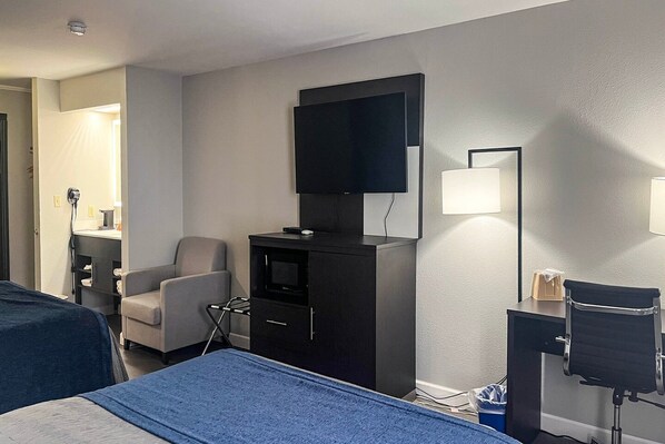 Habitación de hotel con cama, escritorio, silla, televisor y lámpara de pared.