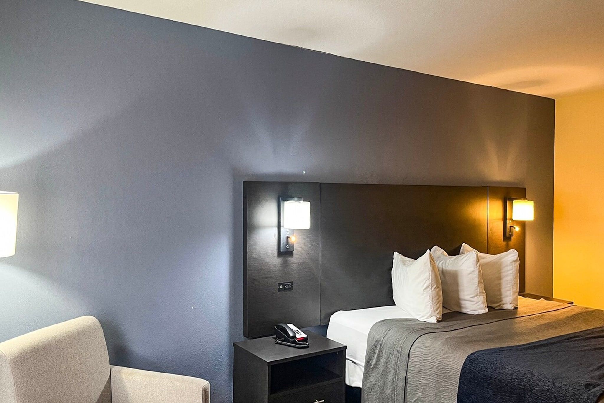 Et hotelværelse med seng, natbord, lampe og en stol.