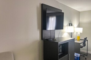 A hotel room with a flat-screen TV mounted on a cabinet, a desk with a lamp, and a trash bin.