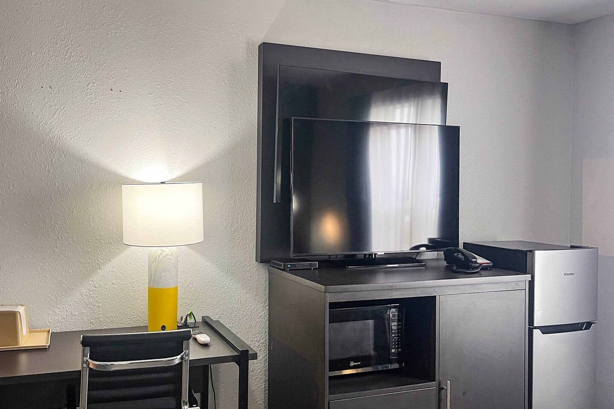 Hotellihuone, jossa on seinälle asennettu taulutelevisio, työpöytä lampun kanssa ja minibaari.