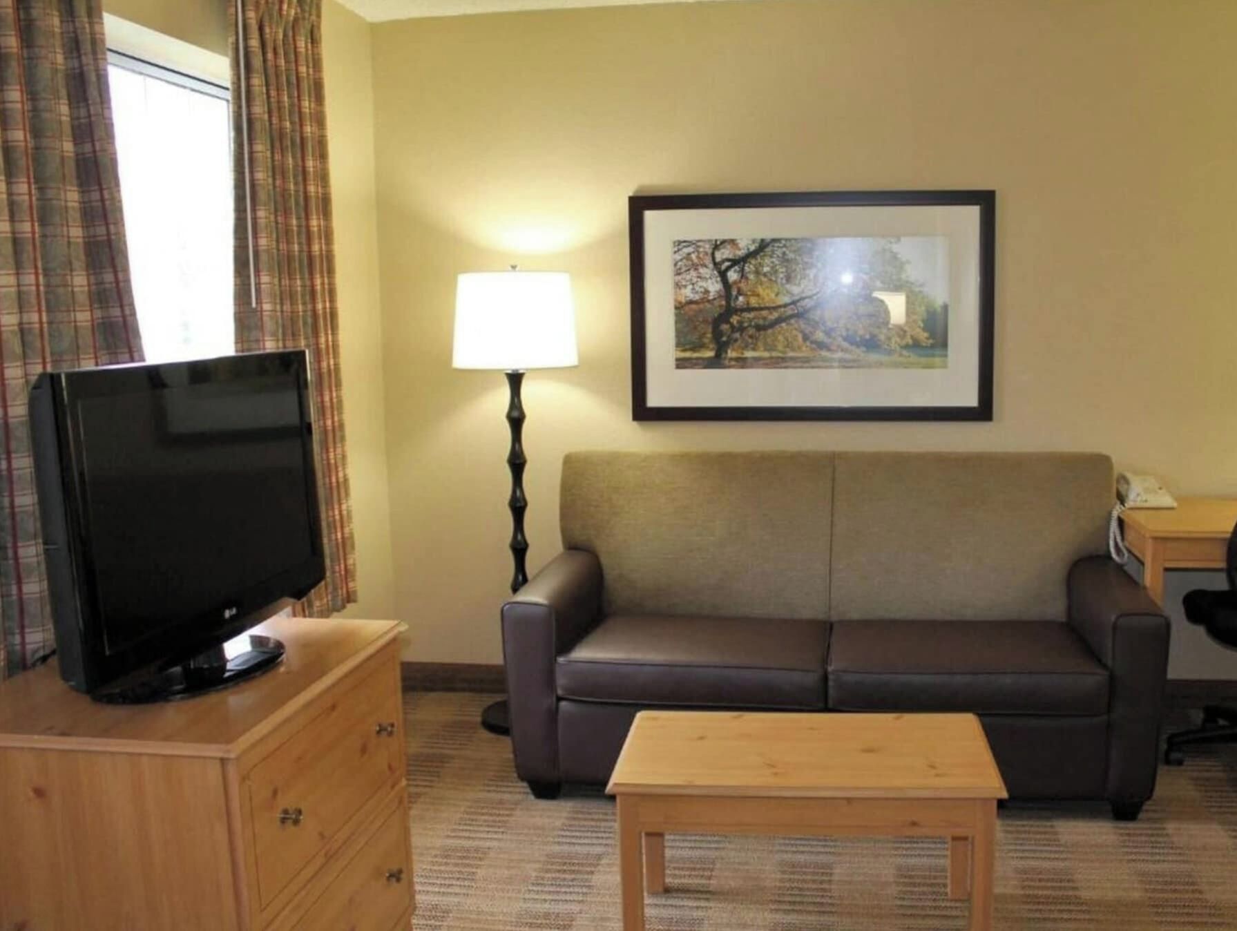 Et hotelværelse med et fladskærms TV på en trækommode, en sofa, et lille træbord, en lampe og et billede i ramme på væggen.