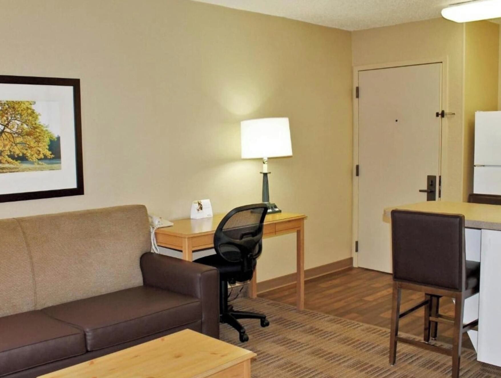 A hotel room with a brown leather sofa, a wooden desk with a lamp, a black office chair, and a door.