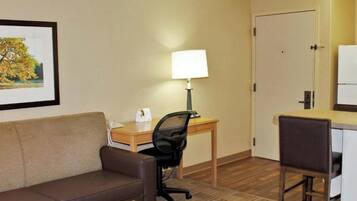 A hotel room with a brown leather sofa, a wooden desk with a lamp, a black office chair, and a door.