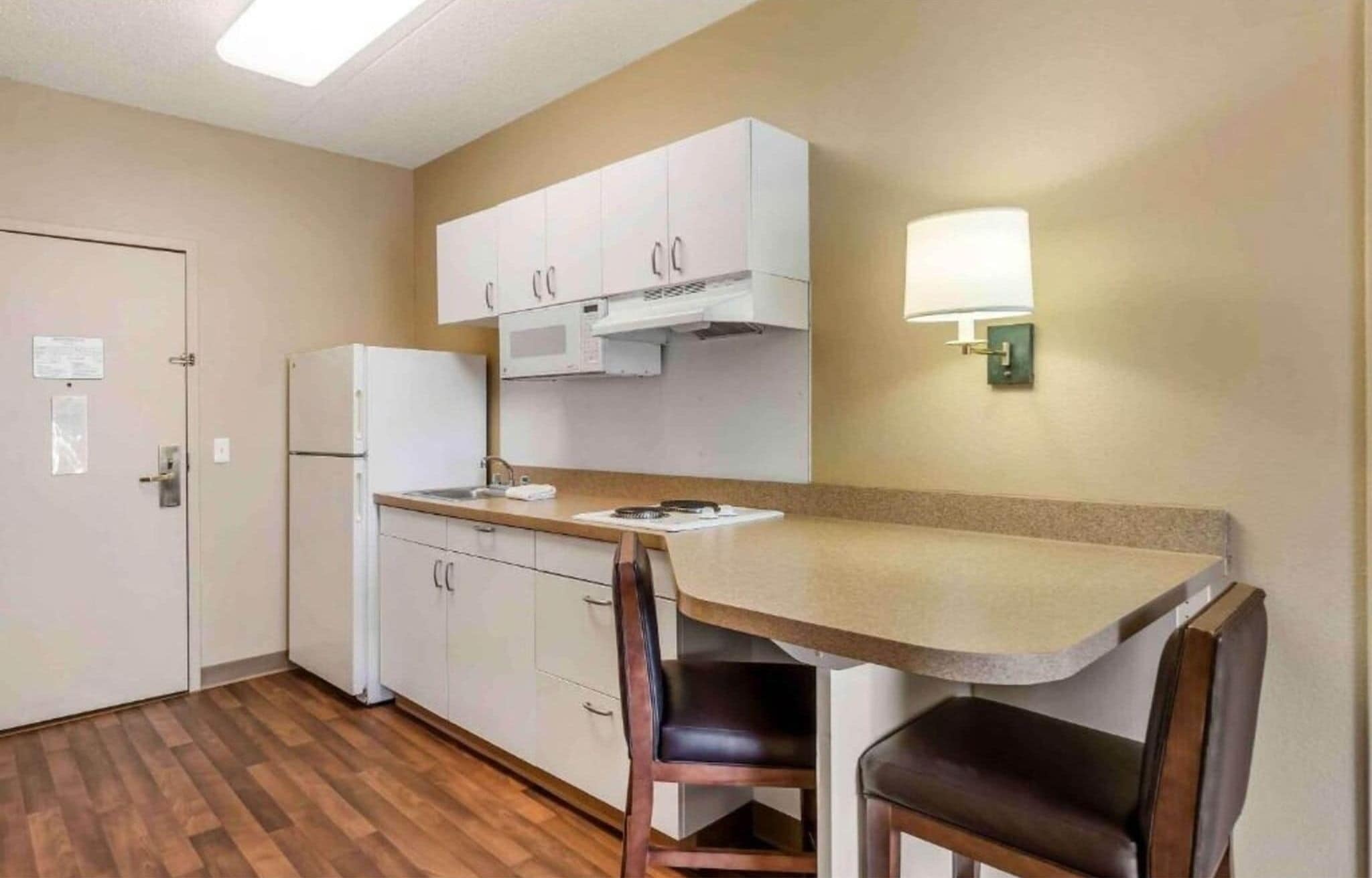 A compact kitchen with a refrigerator, microwave, and a small sink. There are two chairs and a table with a lamp.