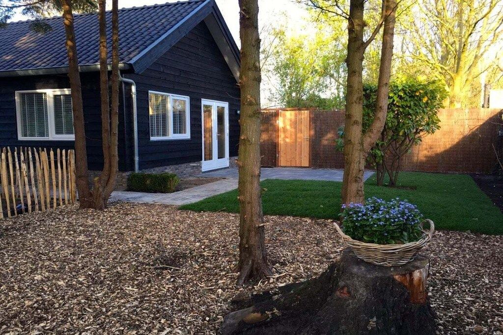 A residential property with a dark-colored house, a wooden fence, a lawn, and a tree stump with a basket of flowers.