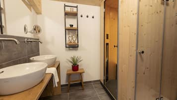 A bathroom with two sinks, a mirror, a towel rack, a small stool with a plant, and a shower enclosure.