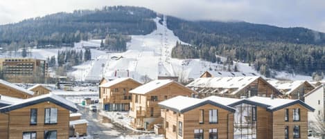 A snow-covered village with wooden buildings, a ski slope in the background, and a clear sky.