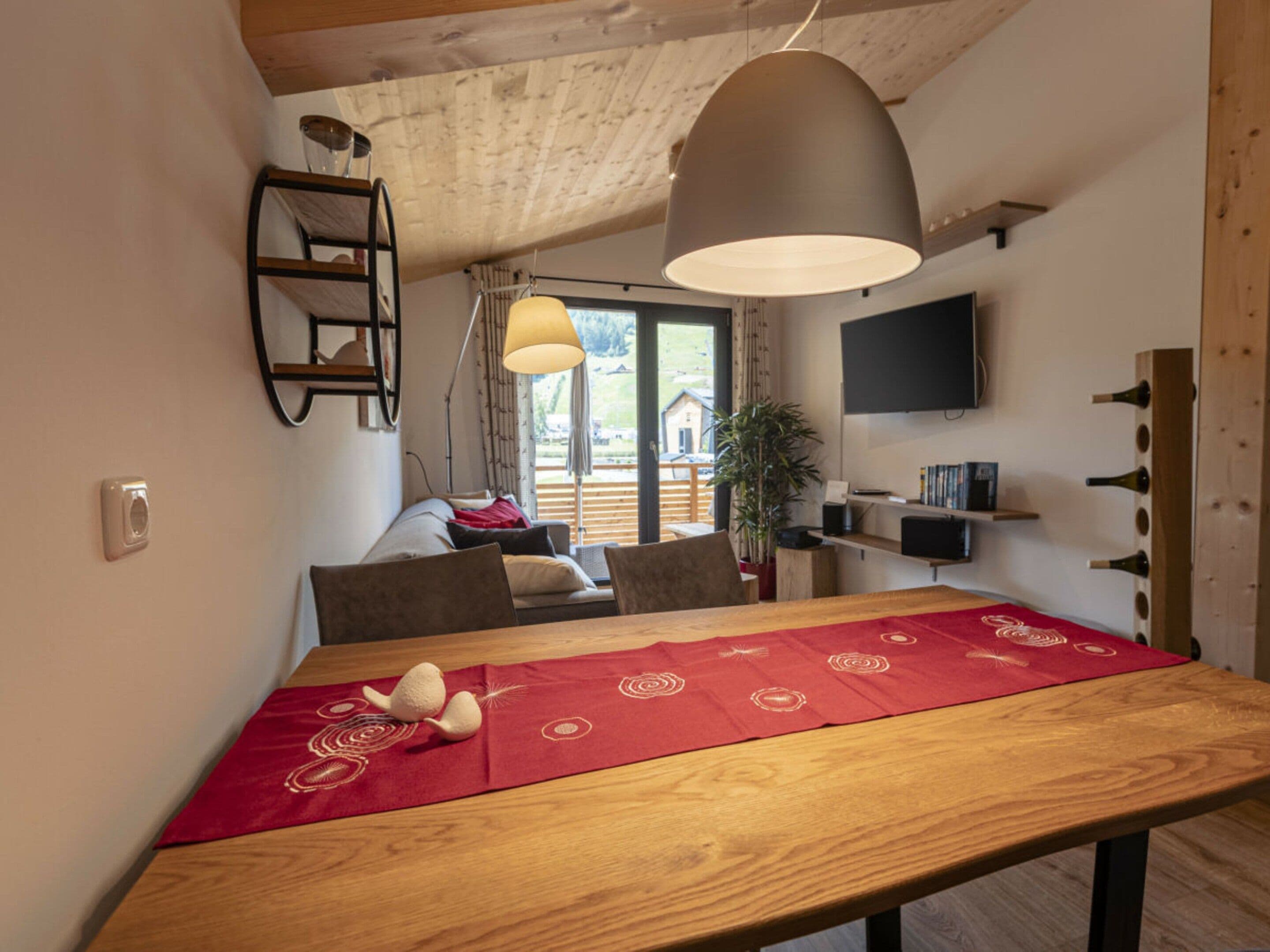 Uma área de refeições com mesa de madeira, um jogo de mesa vermelho, luminária pendente e televisão fixada na parede.