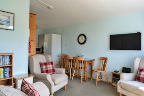 A cozy living room with a sofa, armchairs, a dining table with chairs, a bookshelf, a wall-mounted clock, and a flat-screen TV. - Foxgloves Bungalow (Welcombe)