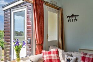 A cozy room with a wooden exterior, a sliding glass door, a red curtain, a fish wall decoration, and a vase with purple flowers. - Foxgloves Bungalow (Welcombe)