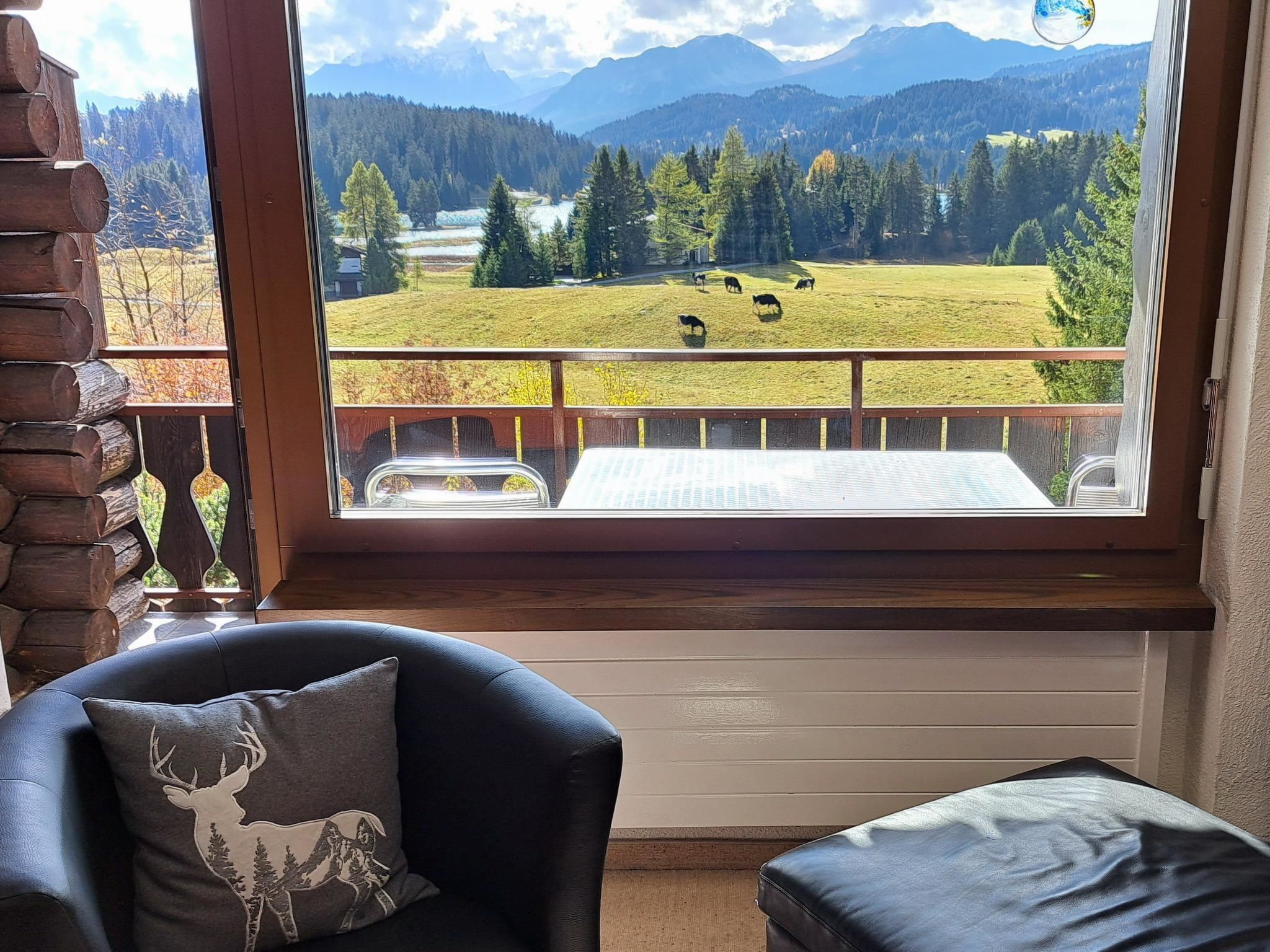 A room with a large window overlooking a green field with grazing animals, a wooden deck, and a leather chair with a decorative pillow.