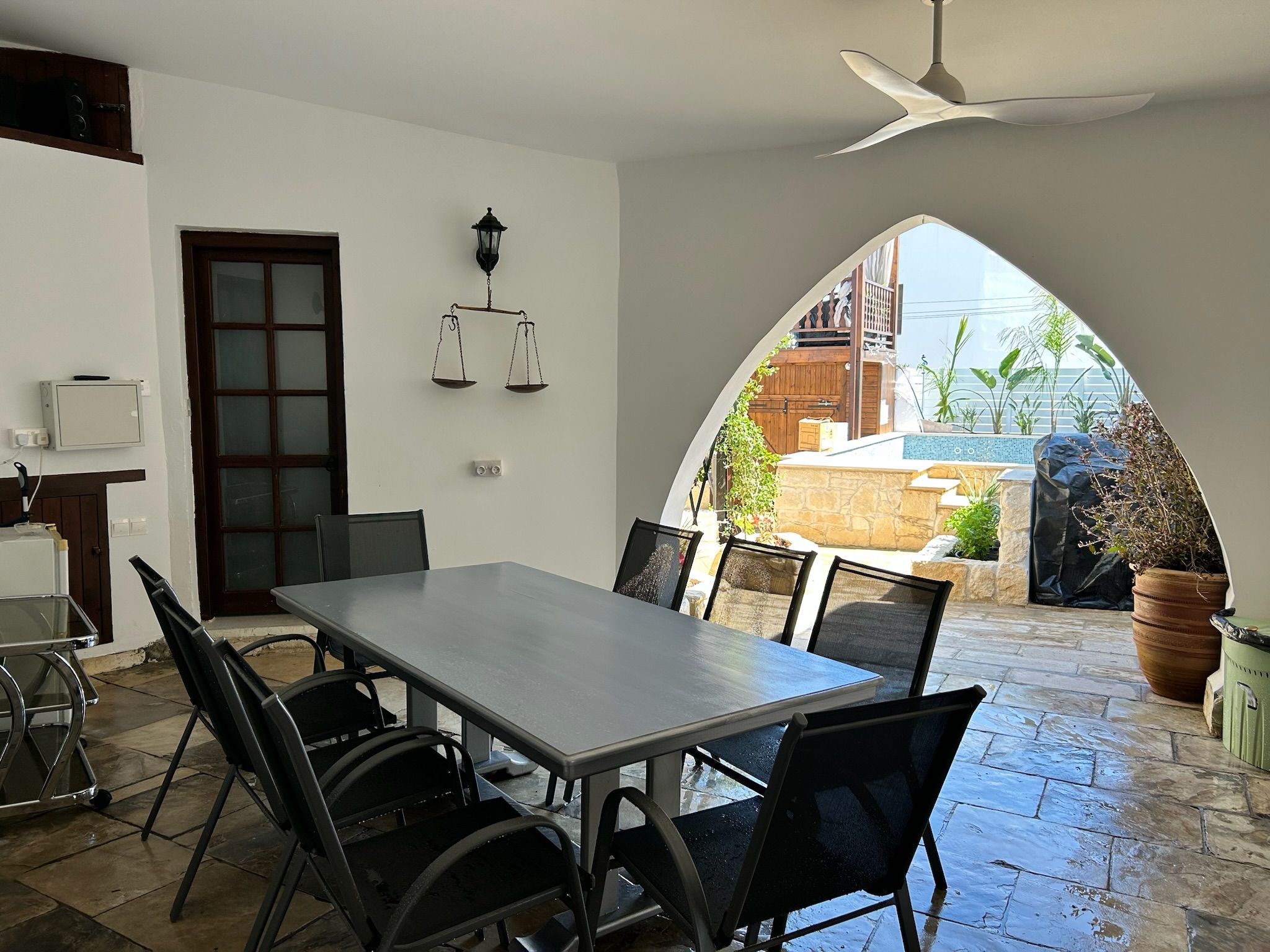 A dining area with a table and chairs, a door, a scale, and a view through an archway to an outdoor space with plants and a pool.