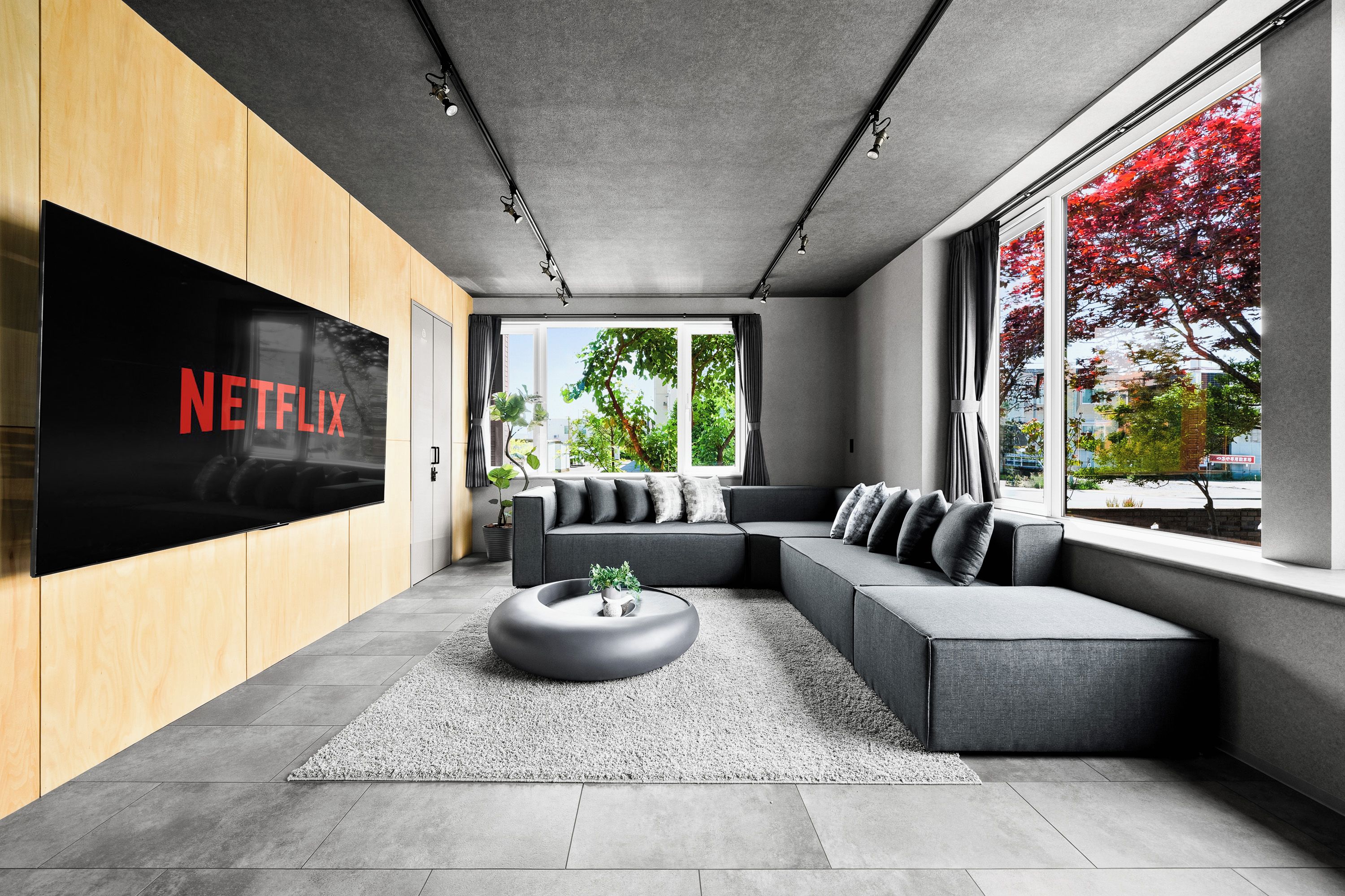Uma sala de estar moderna com sofá seccional, TV de tela plana presa a uma parede de madeira e janelas amplas com vista para árvores.