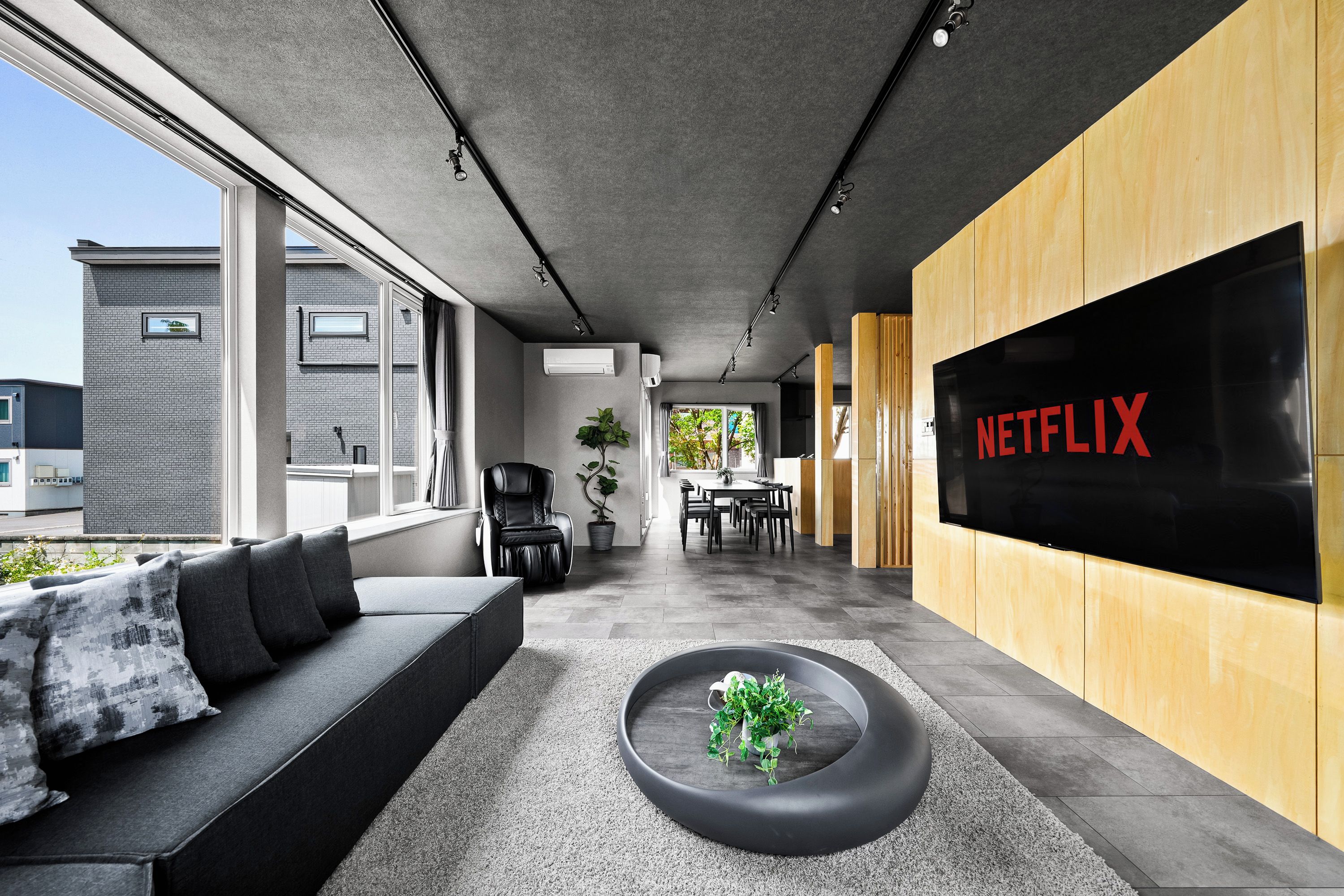A modern interior with a sofa, a coffee table with a plant, a dining area, and a large screen displaying 'NETFLIX'.