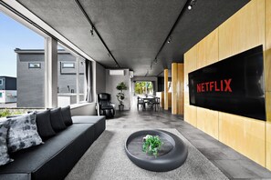 A modern interior with a sofa, a coffee table with a plant, a dining area, and a large screen displaying 'NETFLIX'. - THE CROCUS ASAHIKAWA (Asahikawa)