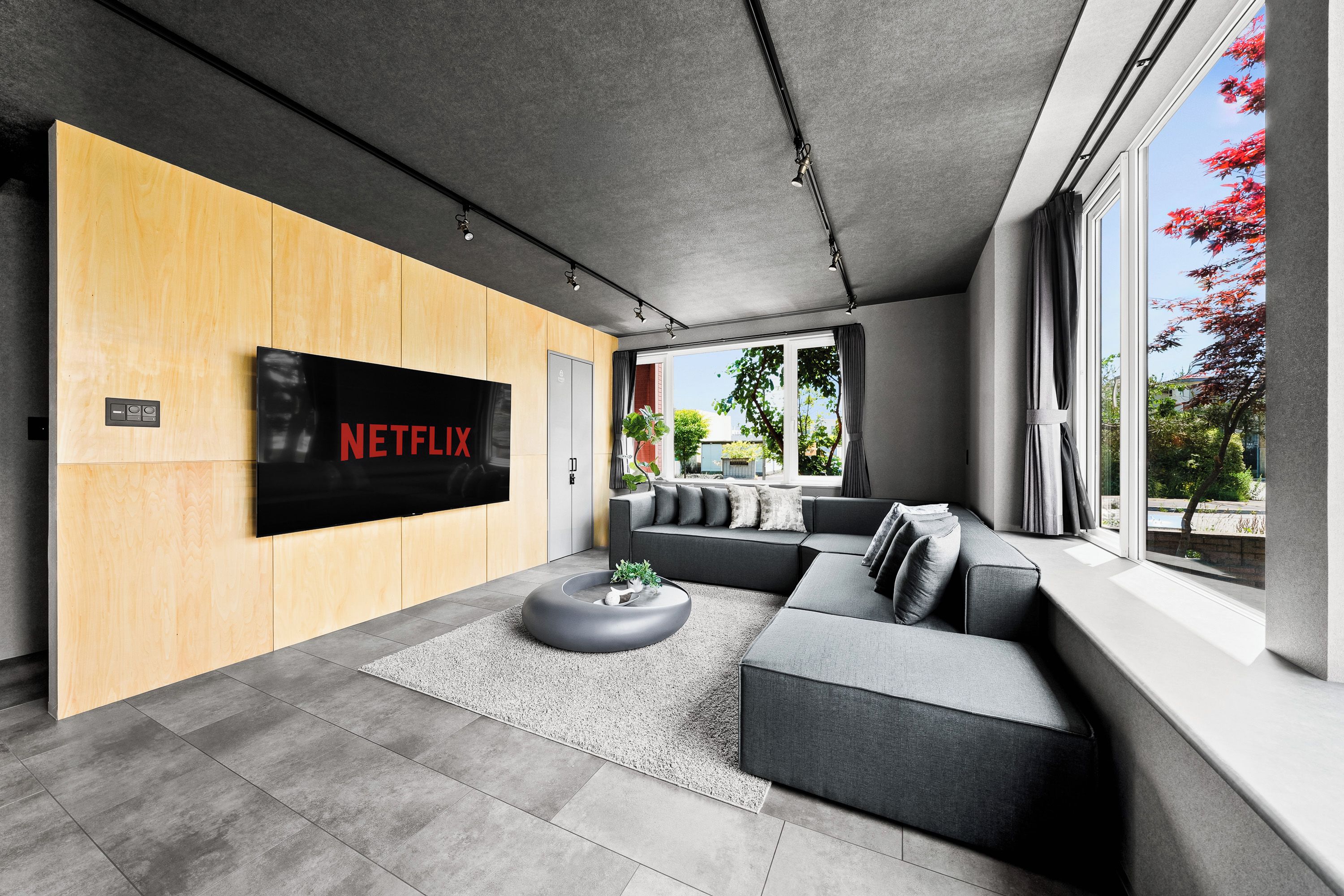 A modern living room with a large flat-screen TV mounted on a wooden wall, grey sofas, a round coffee table, and a large window with a view of trees and a clear sky.