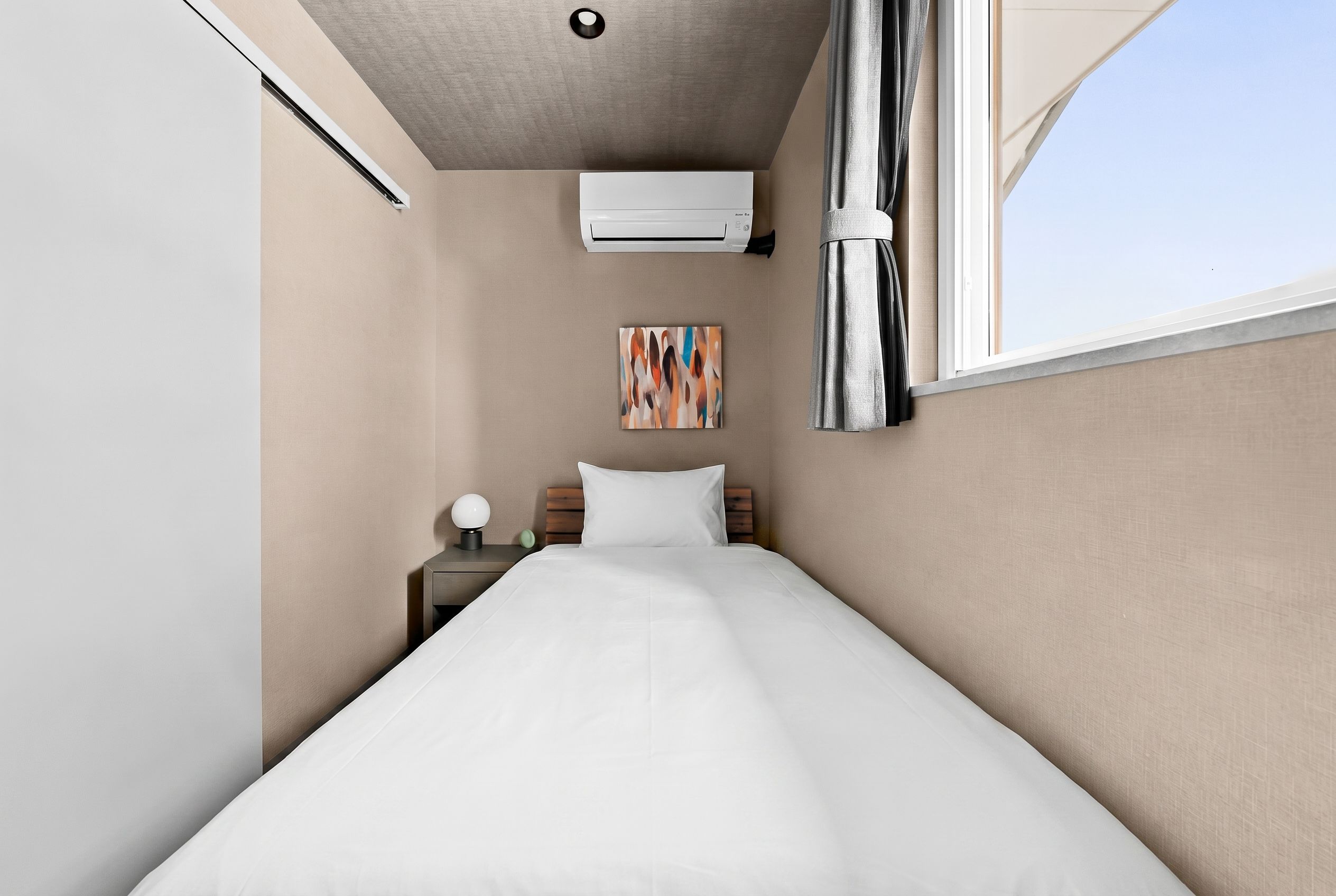 A hotel room with a bed, a nightstand, a wall-mounted air conditioner, a painting, and a window with curtains.