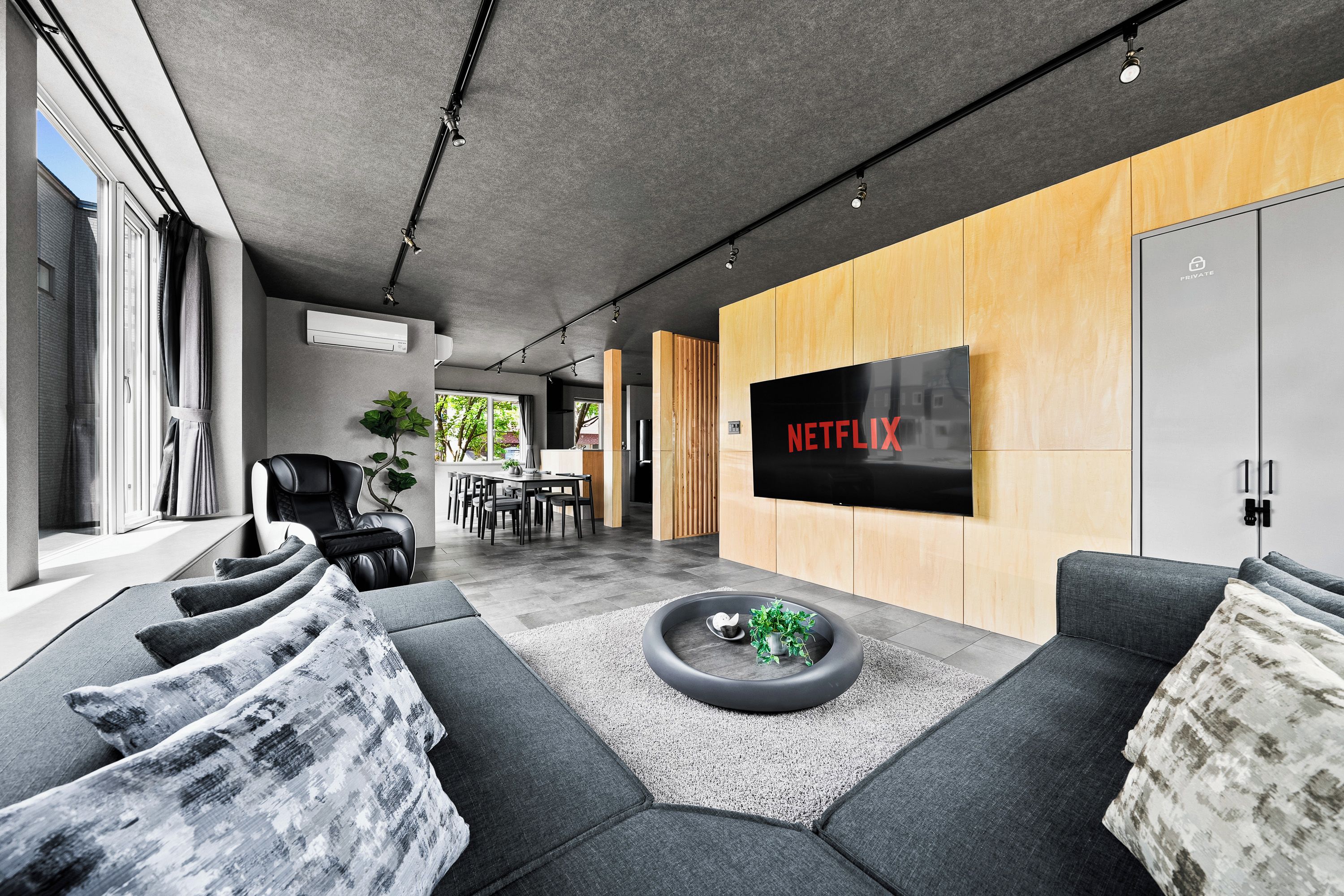 Een moderne woonkamer met een grote flatscreen televisie aan een houten wand, een grijze bank, een salontafel met een plant en een massagestoel.