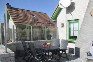An outdoor patio with a table and chairs, a glass enclosure, and a building with a tiled roof and green window frames. - Holiday home with 1 bedroom (Julianadorp aan Zee)