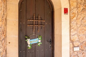Une porte en bois avec un loquet métallique en forme de croix, une pancarte d’accueil ornée de fleurs et une affiche indiquant que les lieux sont surveillés 24 heures sur 24.