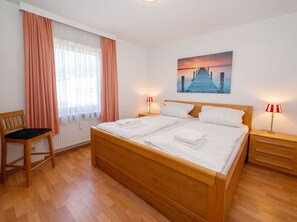 A hotel room with a wooden bed, two bedside tables with lamps, a chair, a window with curtains, and a painting on the wall. - Stunning Vacation Rental No. 32 at Haus Schloss am Meer in a Central Location (Büsum)
