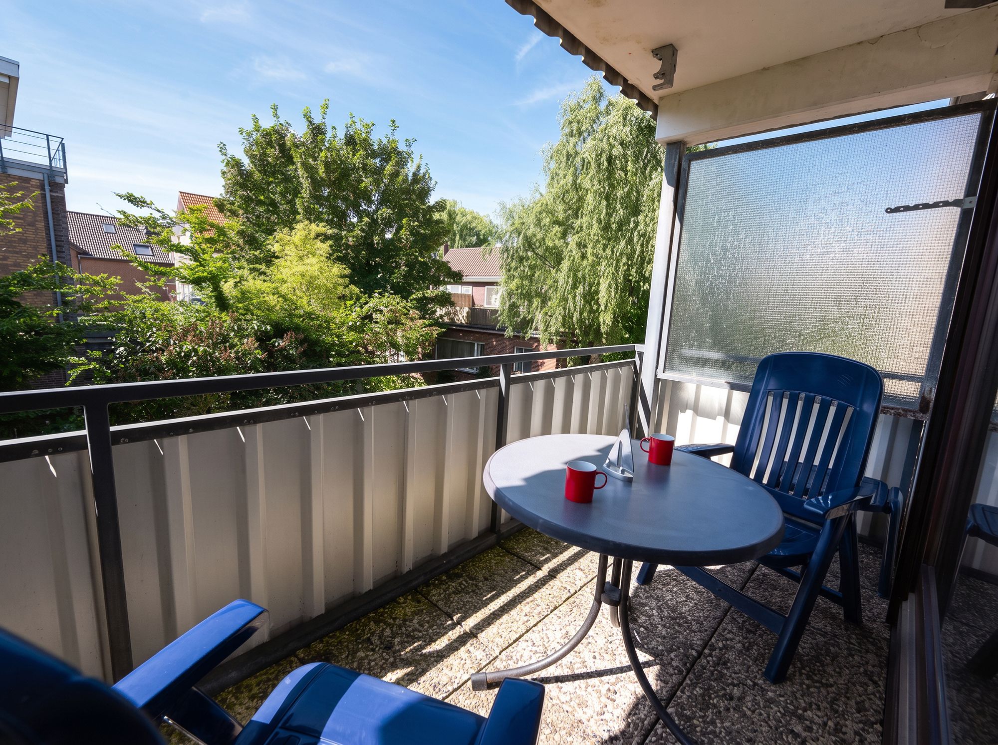A balcony with a table and chairs, a red mug, and a view of trees and buildings.