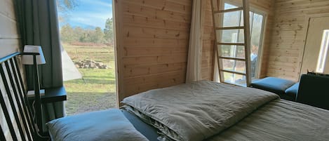 Une pièce de cabane en bois comprenant un lit, une fenêtre et donnant sur un champ verdoyant.