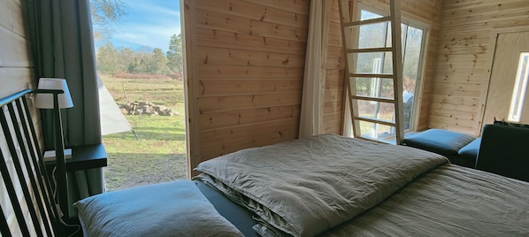 A wooden cabin room with a bed, a window, and a view of a grassy field.