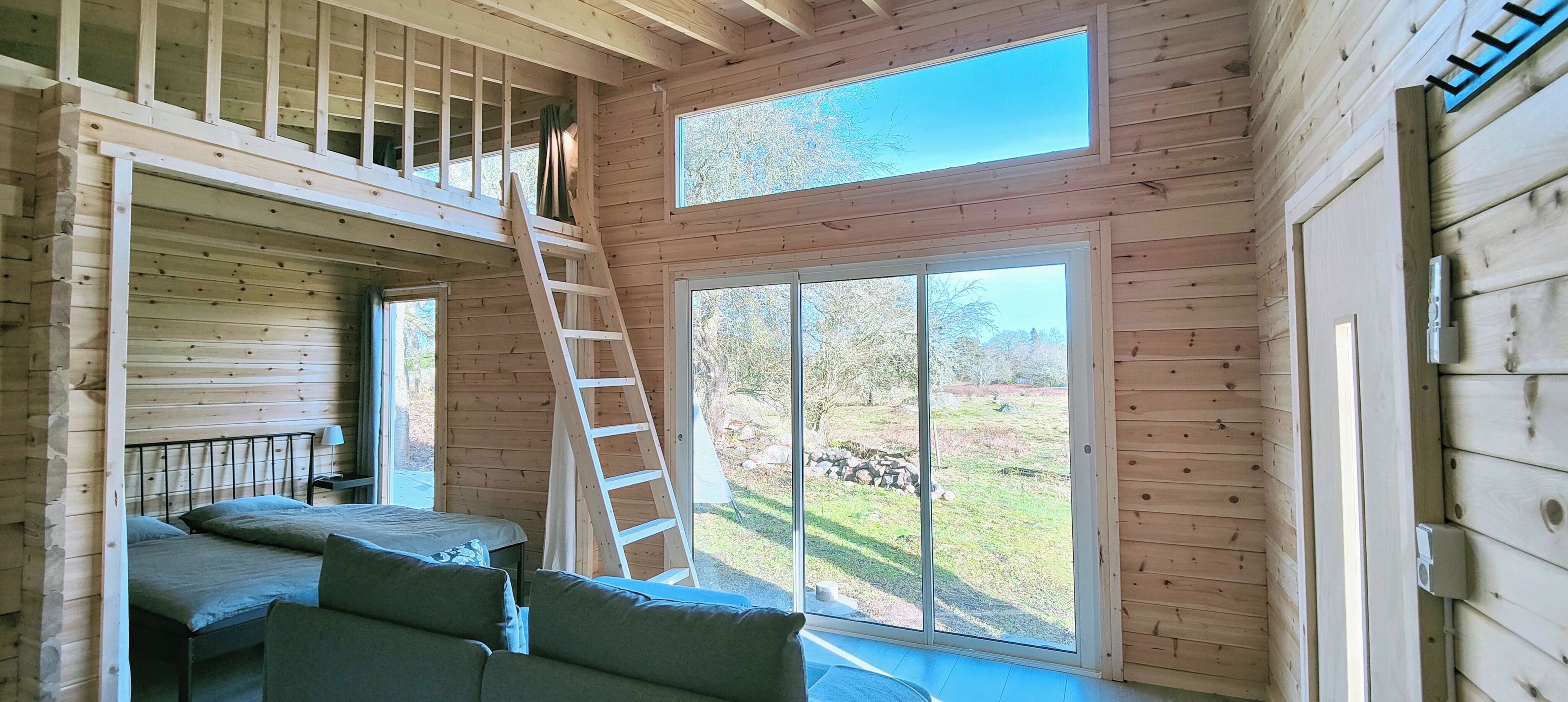 A cabin interior with a loft bedroom, a sofa, a ladder, and large windows.