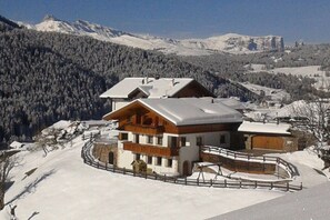 A chalet-style building with a snow-covered roof, surrounded by a wooden fence, set against a backdrop of snow-covered mountains and a clear sky.