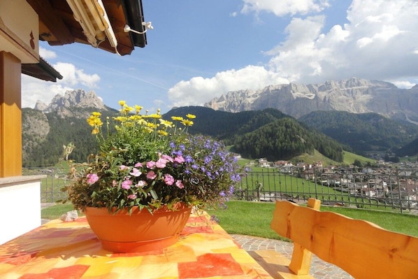 A balcony with a potted plant and a wooden bench, overlooking a mountainous landscape and a town.