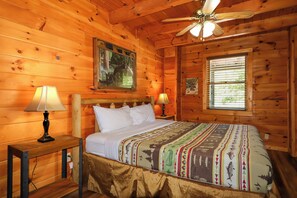Ein Zimmer einer Holzhütte mit einem Bett, zwei Nachttischlampen, einem Deckenventilator und einem Fenster mit Jalousien.