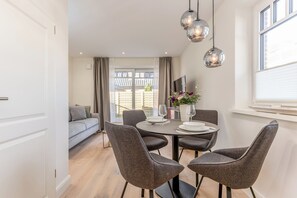 Uma área de refeições com mesa posta para quatro pessoas, cadeiras estofadas cinzas, um vaso com flores e uma luminária pendente moderna.