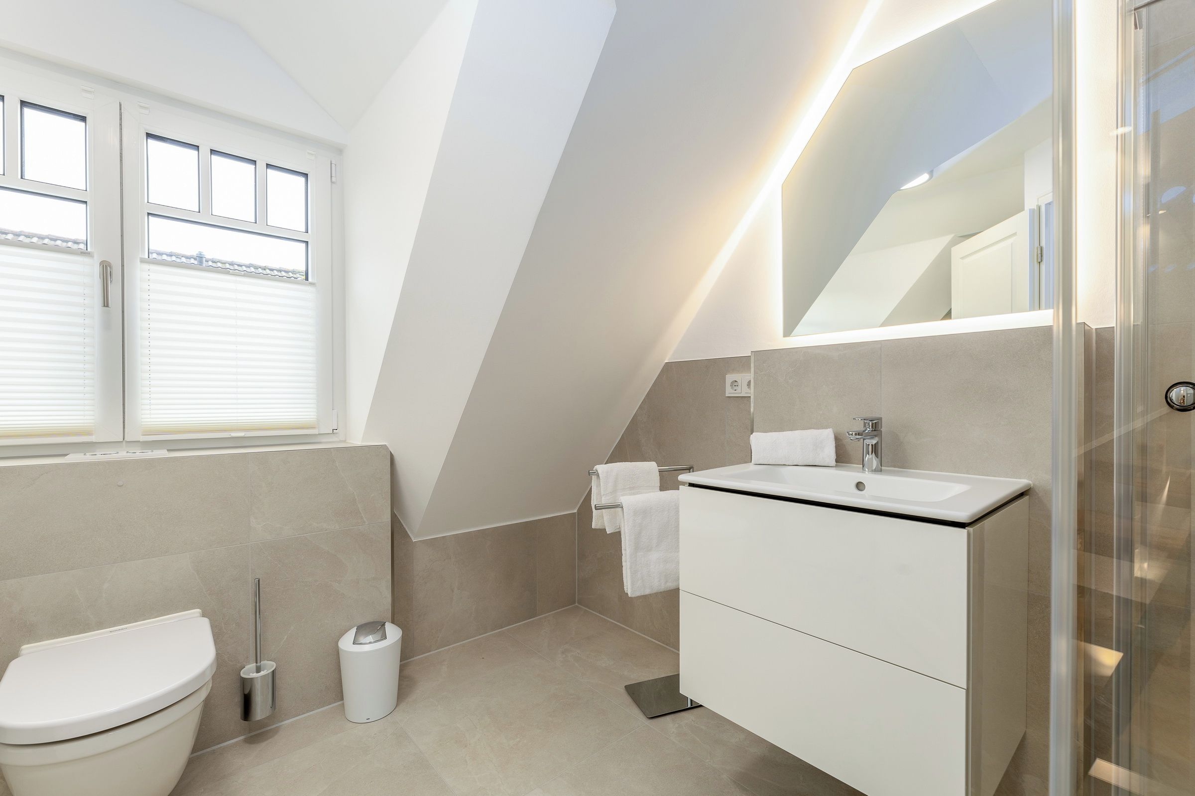 A modern bathroom with a shower, toilet, and sink, featuring a sloped ceiling and large windows.
