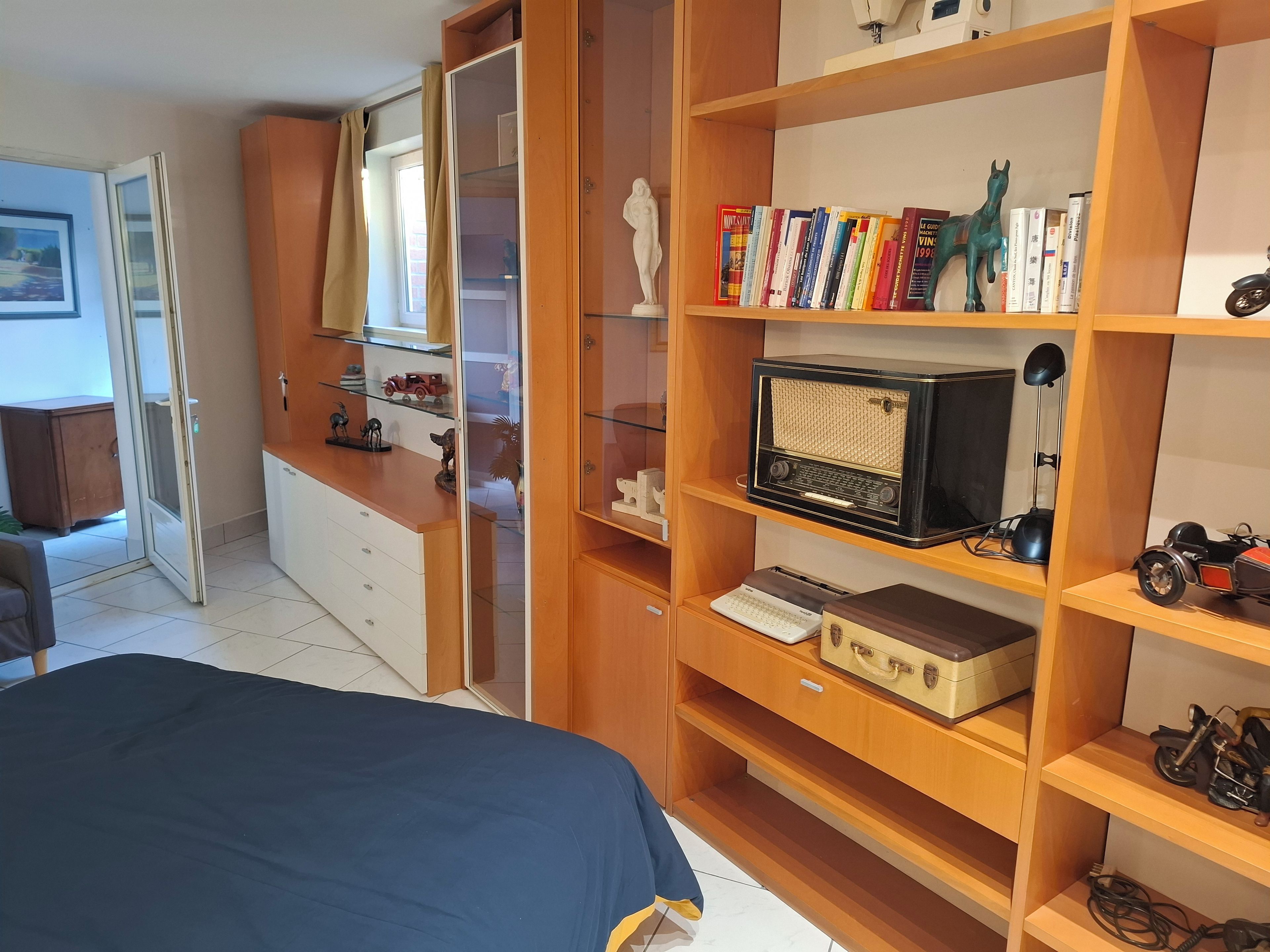 Un dormitori amb un llit, un armari de fusta, prestatges amb llibres i un aparell de ràdio, i una taula de treball amb una làmpada.