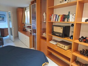 Een slaapkamer met een bed, een houten garderobe, planken met boeken en een radio, en een bureau met een lamp.