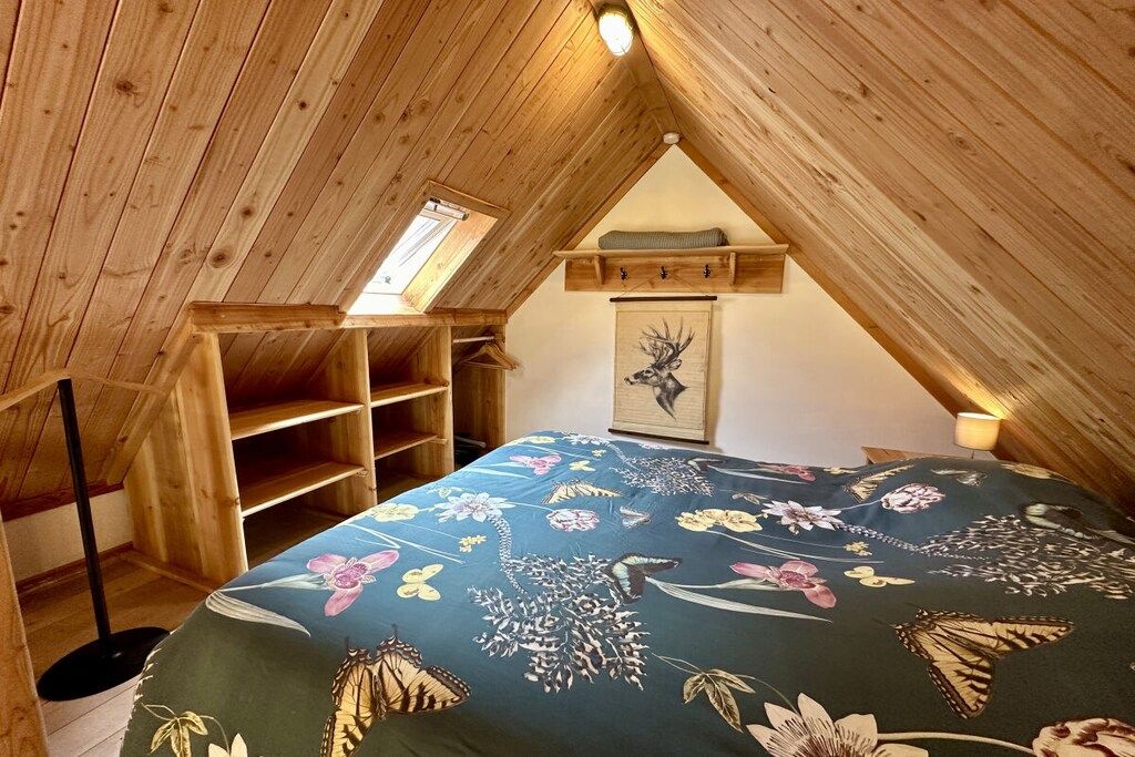 Une chambre avec un plafond en pente, des poutres en bois, un puits de lumière, un lit recouvert d’une courtepointe à motifs floraux, une armoire en bois et un tableau encadré accroché au mur.
