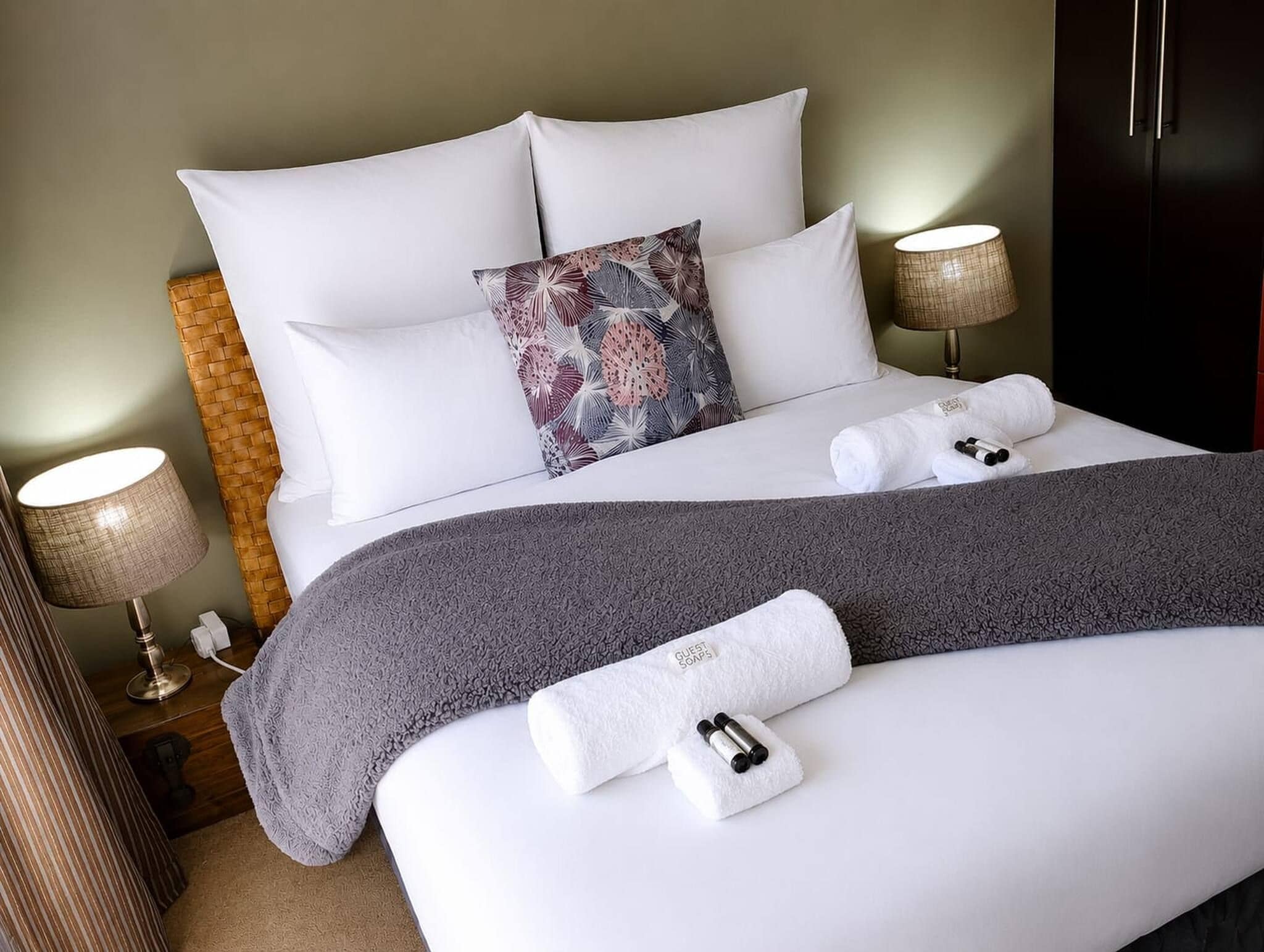 A hotel room with a neatly made bed, a decorative pillow, a grey blanket, and a set of toiletries on the bed.