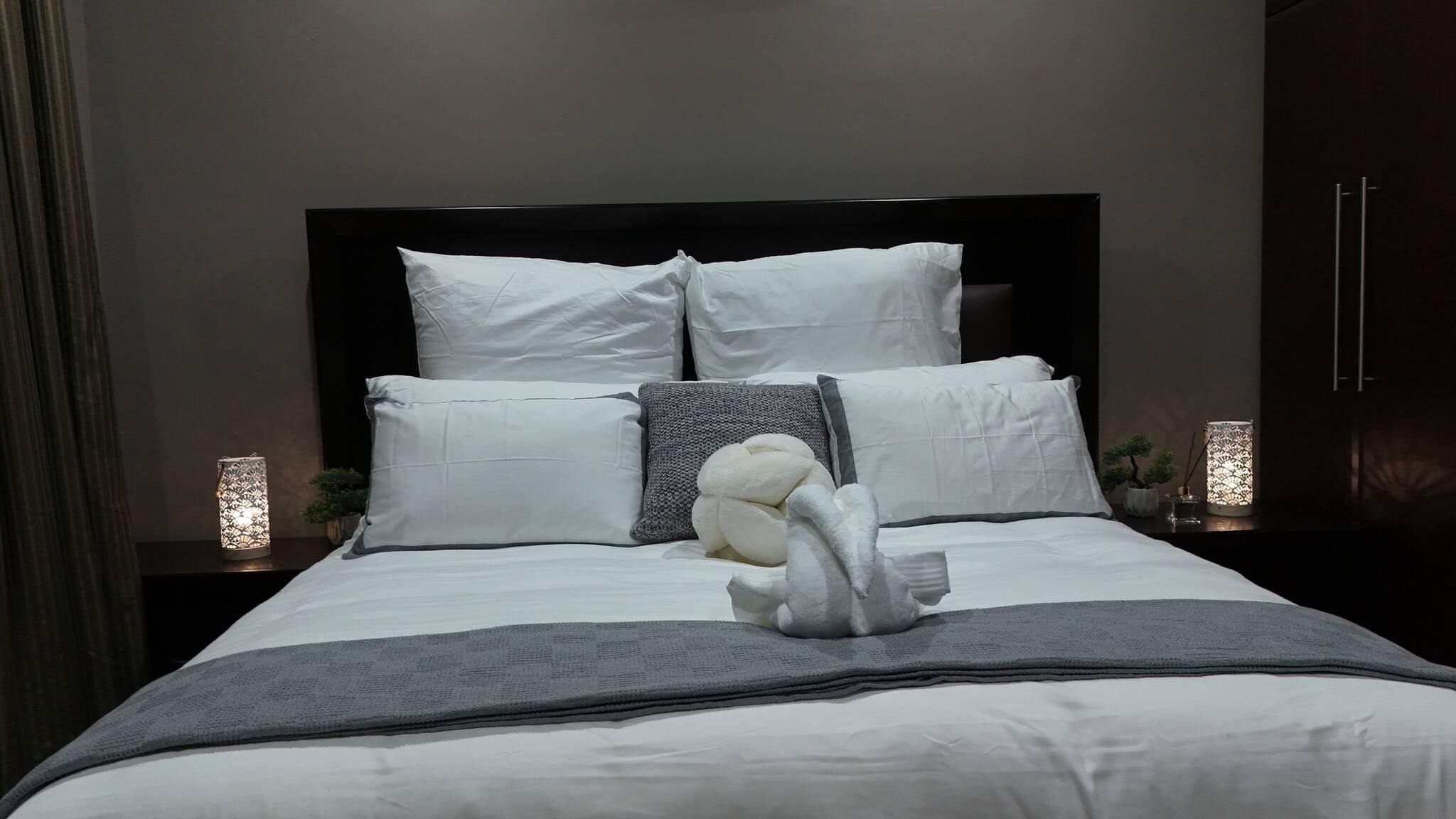 A neatly made bed with white linens and a gray throw blanket, flanked by two bedside lamps and a nightstand with a potted plant.