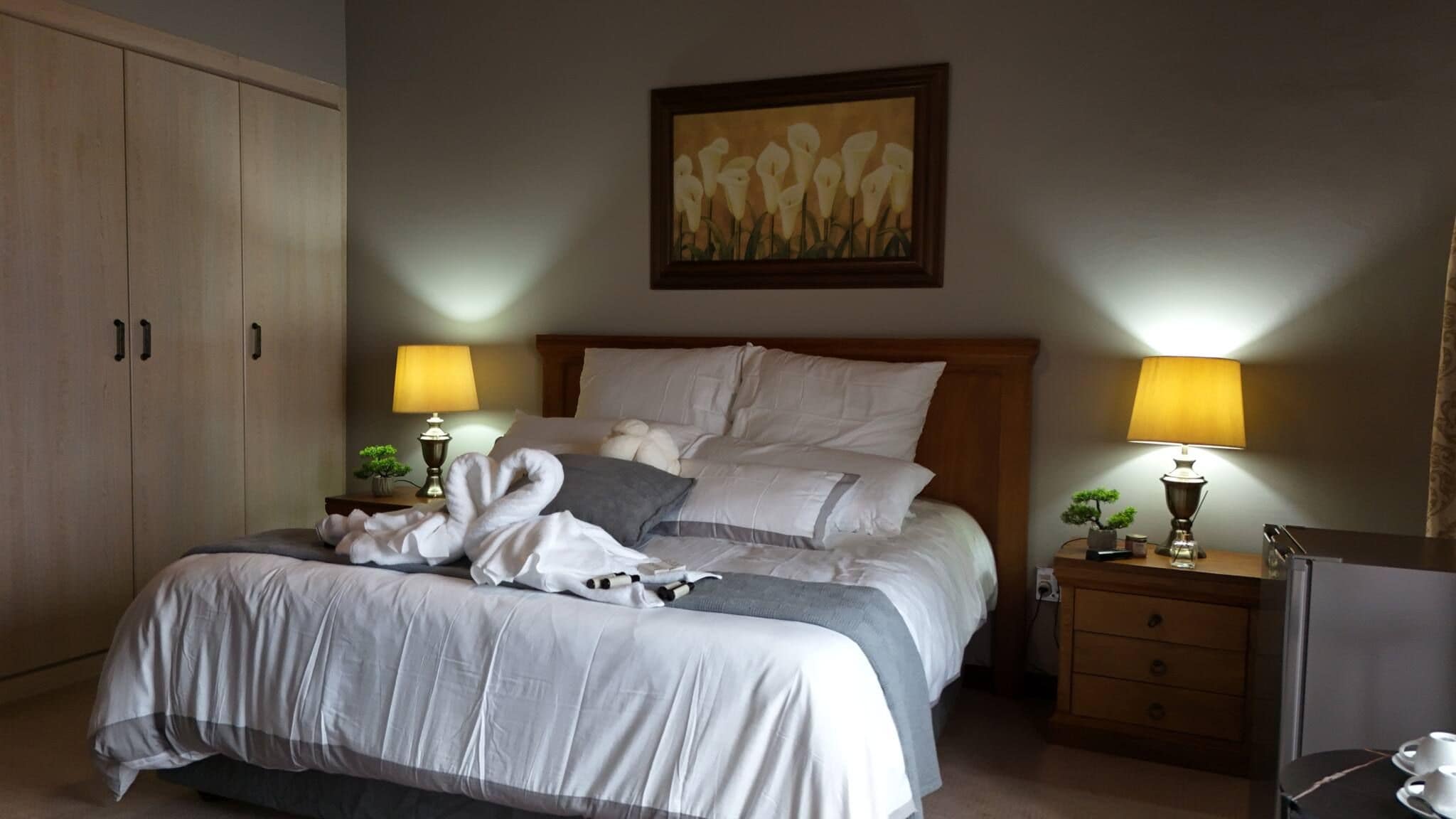 A hotel room with a bed, two bedside lamps, a painting, a wardrobe, and a desk with a lamp.