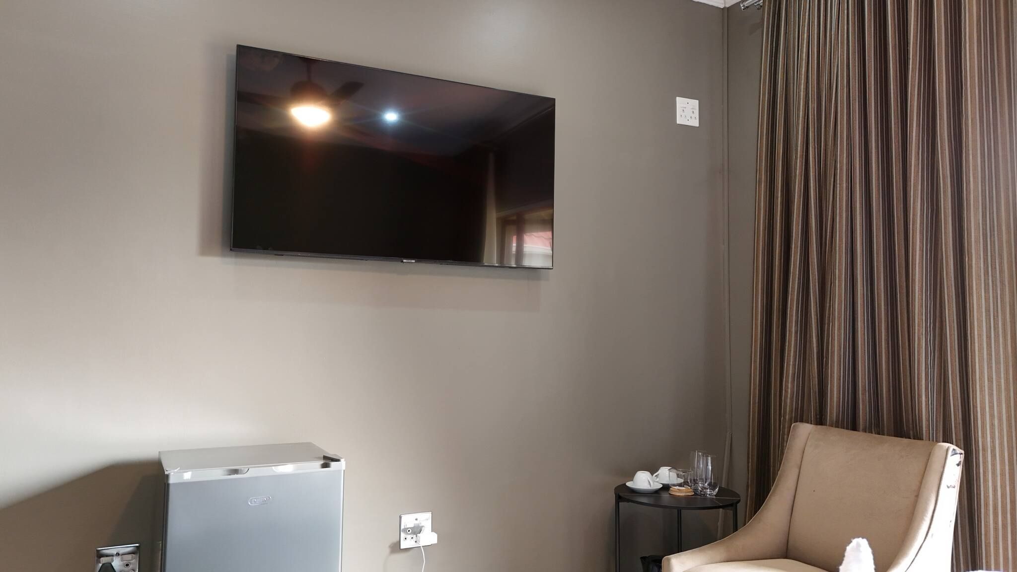 Quarto com televisão de tela plana na parede, uma pequena mesa com cadeira e um refrigerador.