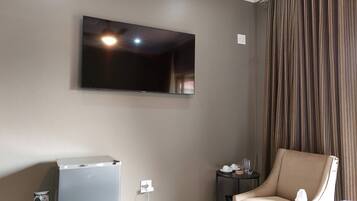 Quarto com televisão de tela plana na parede, uma pequena mesa com cadeira e um refrigerador.