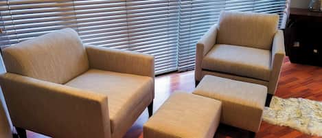 A room with two armchairs and a matching ottoman, a wooden floor, and a window with blinds.
