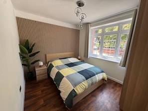 A bedroom with a wooden slat wall, a bed with a checkered bedspread, a nightstand, a potted plant, and a window with a view of the outside.