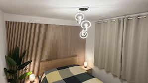 Quarto com parede de ripas de madeira, cama com colcha xadrez, abajur e luminária pendente.
