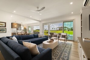 A modern living room with a large sofa, a coffee table, and a view of a golf course through the glass doors. - Hale Olina by Alekona Kauai (Koloa)