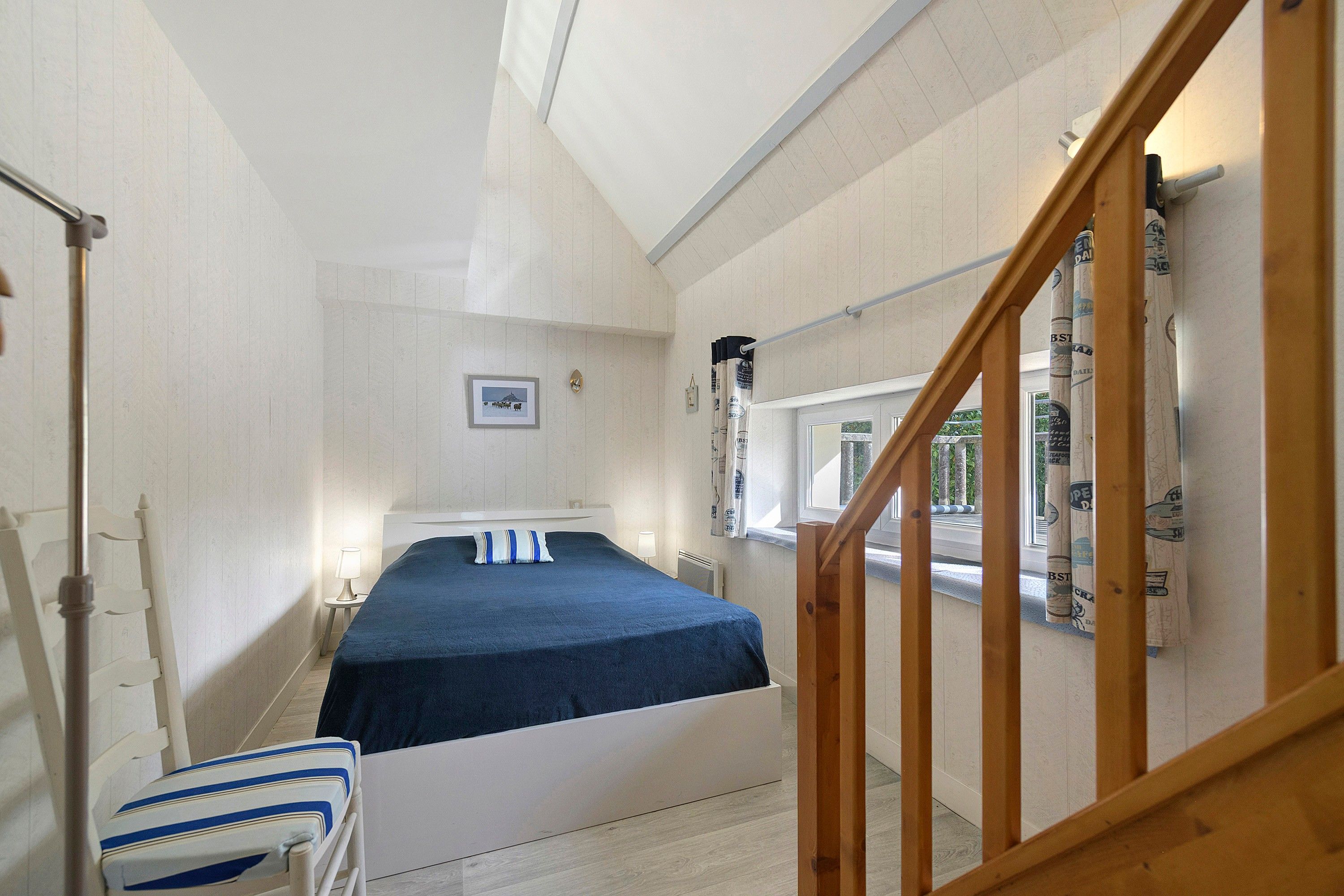 Ein Schlafzimmer mit einem Bett, einem Nachttisch, einem Sessel, einem Fenster mit Vorhängen und einer Holztreppe.