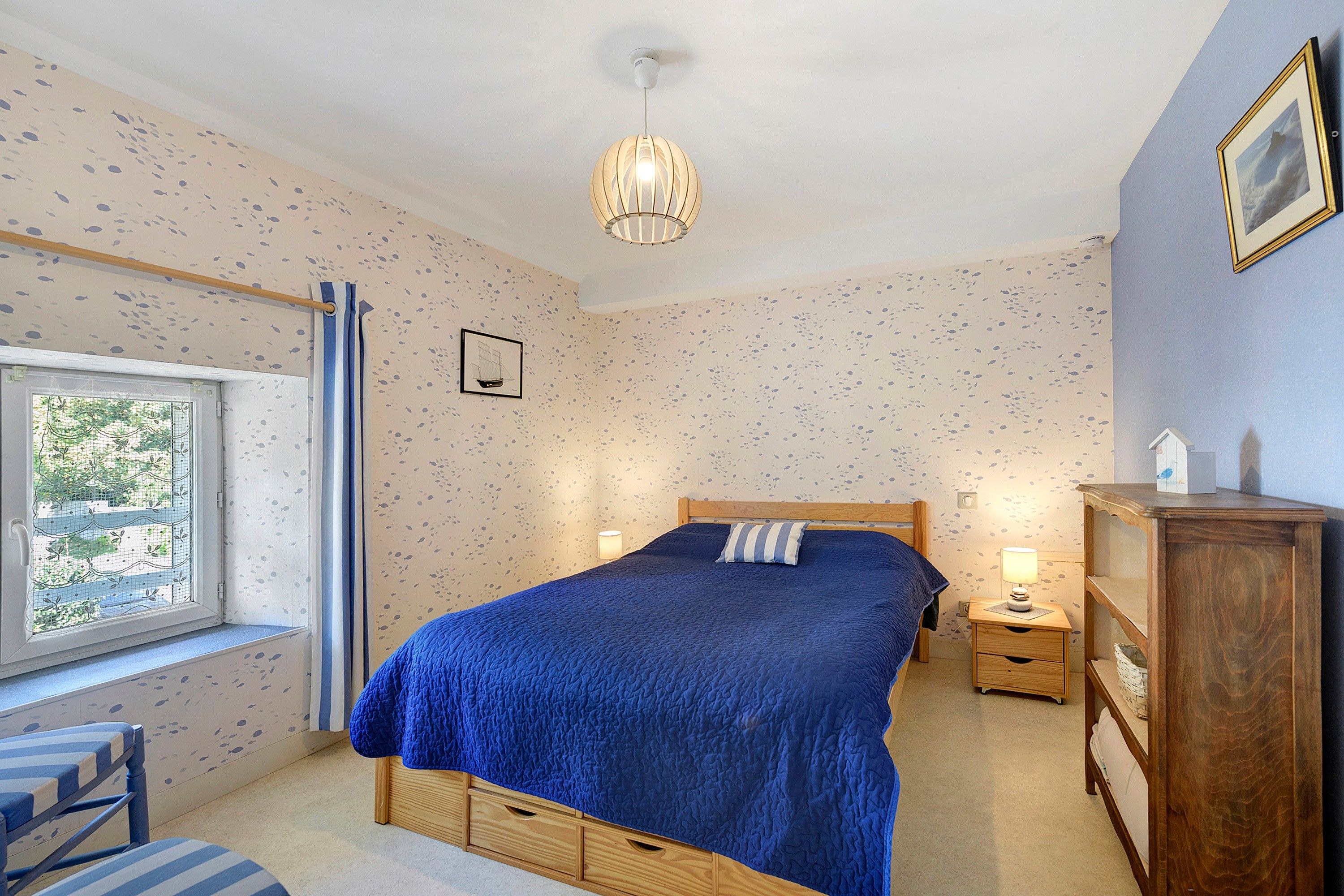 A bedroom with a large bed, a window with curtains, a bedside table with a lamp, and a wooden dresser.