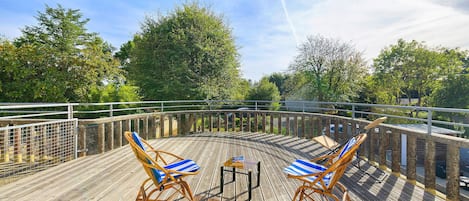 A wooden deck with two wicker chairs and a small table, surrounded by a metal railing and trees.