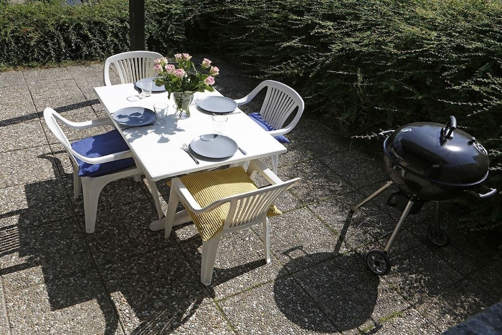 Un espace extérieur aménagé pour les repas, avec une table blanche dressée pour quatre personnes, entourée de chaises blanches, d’un vase rempli de fleurs et d’un barbecue.